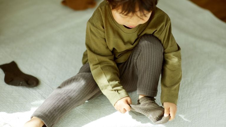 A toddler or preschool-aged child demonstrates independence and fine motor skills while sitting on a quilted floor mat. The child is captured in a candid, top-down view, intently concentrating on pulling a grey non-slip sock onto their foot, illustrating a milestone in developing executive function and self-care.