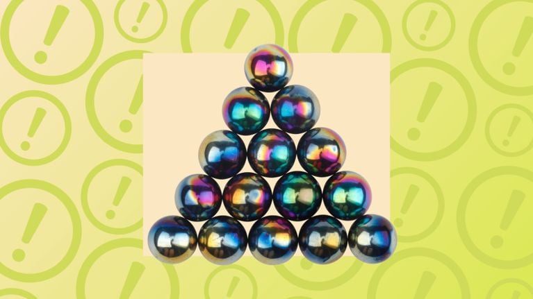 A collection of 15 shiny, rainbow-colored metallic spheres arranged in a perfect triangle. The background features a repeating pattern of light green exclamation point symbols inside circles, signaling a safety alert or product recall related to these high-powered magnetic toys.