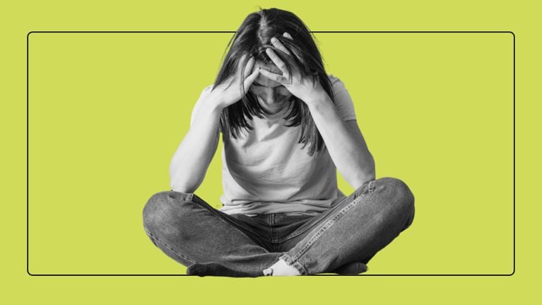 A woman sitting cross-legged in jeans and a t-shirt, holding her head in her hands with a stressed expression.