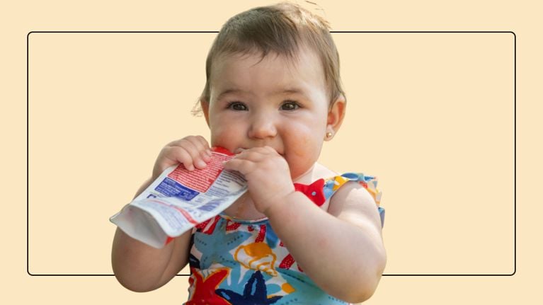 A close-up portrait of a toddler eating from a squeeze pouch with messy hands and face, with a green background.