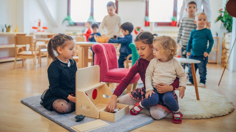 Three young children playing with a wooden Montessori object permanence box on a daycare rug.