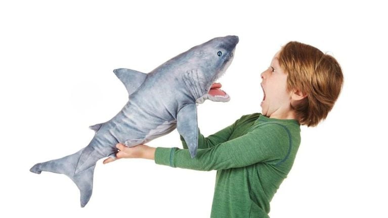 A young boy holds a large, realistic shark plush toy with his mouth open in a playful, surprised expression.