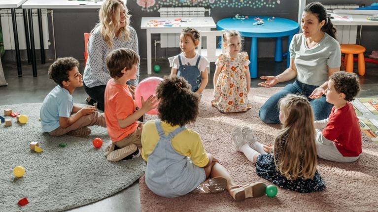 A diverse group of preschoolers and two teachers sitting in a circle on a rug during daycare morning meeting.