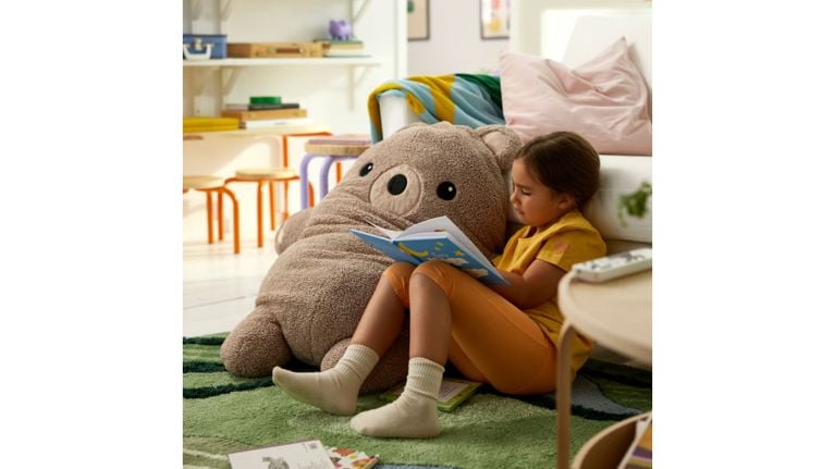 A young girl sits on a green rug, reading a book while leaning against a large, plush brown bear floor cushion.