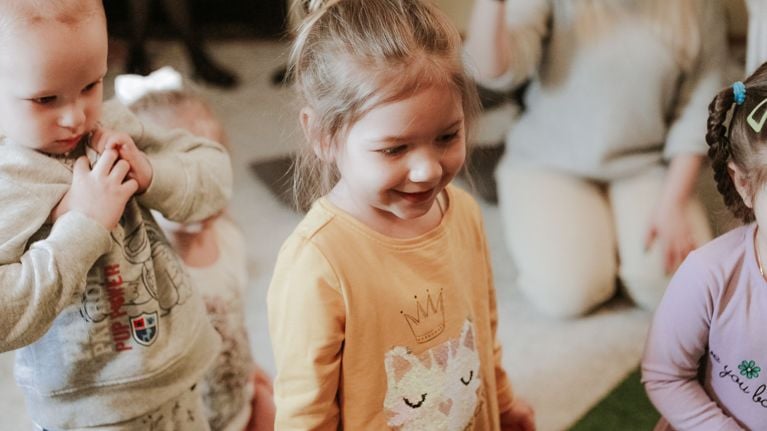 A young girl in a yellow shirt smiling during a daycare morning activity with other children nearby.