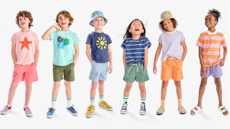 Six diverse children laughing and posing in colorful summer outfits, patterned t-shirts, and bright shorts against a white backdrop.