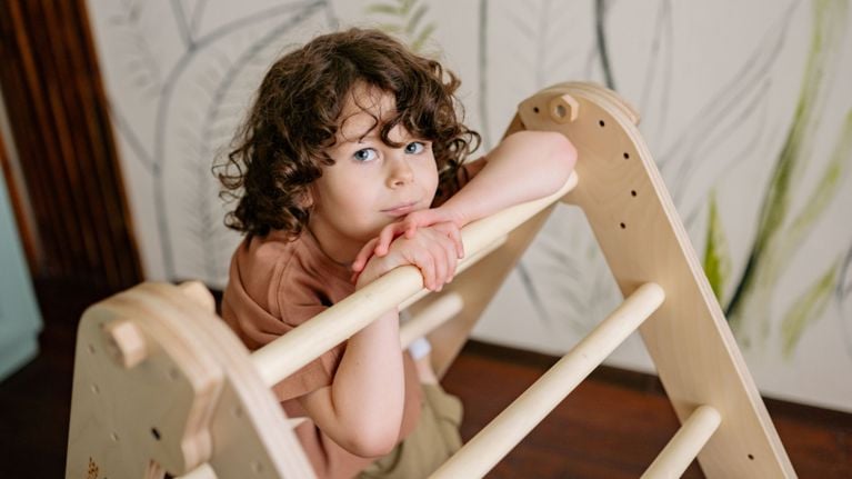 A young child with brown curly hair leaning their chin on a wooden Pikler triangle climbing frame in a playroom.