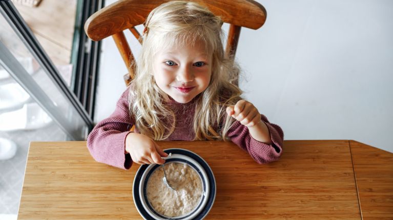 A blonde toddler sitting at a wooden table smiling while eating a bowl of oatmeal.