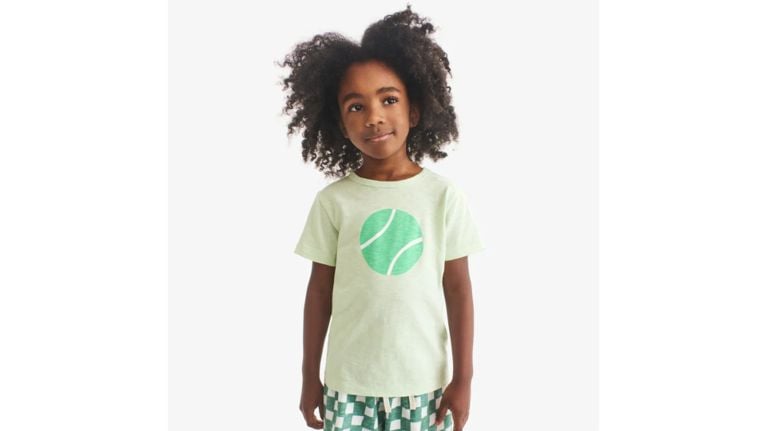 Young child with curly hair wearing a pale green Primary t-shirt with a large tennis ball graphic.