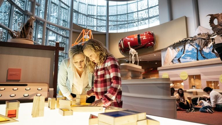 An adult woman with blonde hair and a young girl with curly hair stand close together, looking down at wooden building blocks on a glowing white light table. They appear to be in a large, modern museum hall with a high glass window wall in the background. Around the room, various exhibits are visible, including a stuffed animal display, a red vintage car mounted on a wall, and a large dinosaur skeleton. In the lower right background, other visitors sit on the floor.