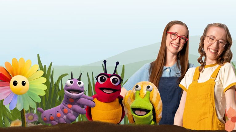 Alt Text Analysis Alt Text (The Code): Two smiling women stand behind colorful garden-themed puppets, including a caterpillar, ladybug, and snail.