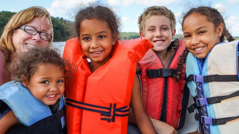 A smiling mother and four children wearing brightly colored life jackets while enjoying a family boat ride on a lake.