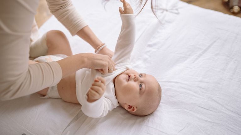 A mother gently dressing her infant baby in a white long-sleeve onesie on a white bed.