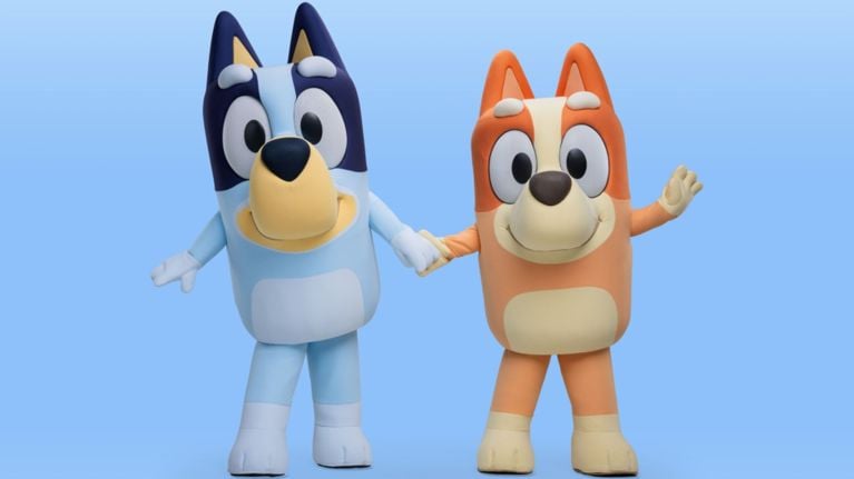 Life-sized Bluey and Bingo mascot costumes holding hands and waving against a blue background.