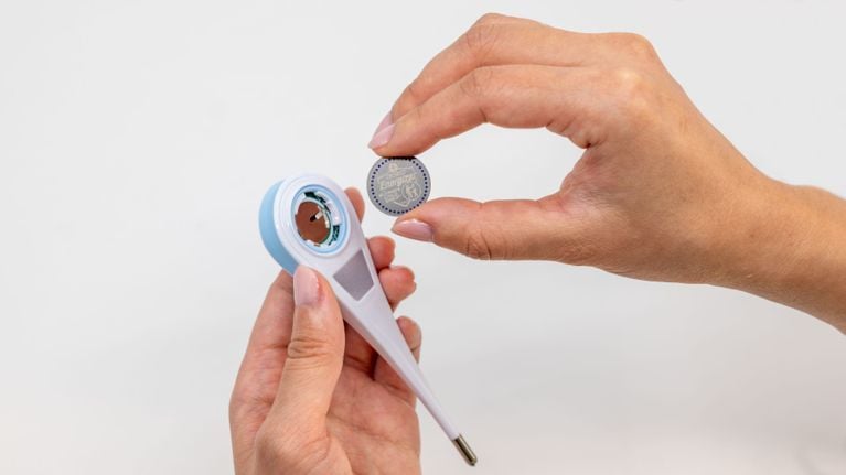 A hand holds an Energizer CR2032 lithium coin battery next to an open digital thermometer against a white background.