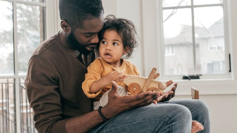 A father cuddles his toddler on his lap while helping them hold a wooden toy airplane in a bright living room.