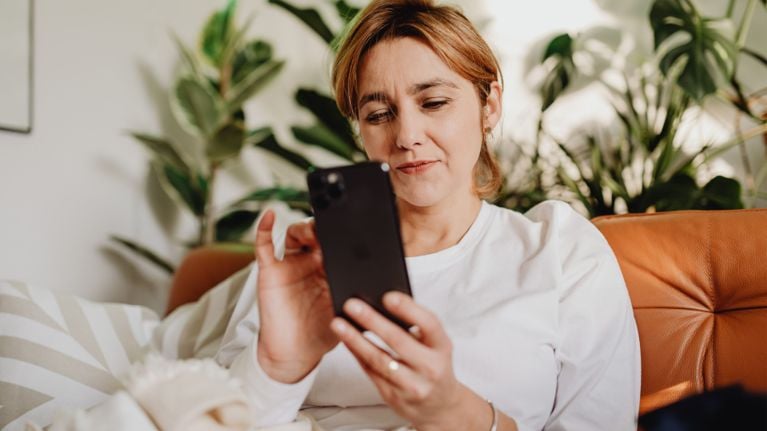 A mother with a focused expression sits on a leather couch, looking at her smartphone at home.