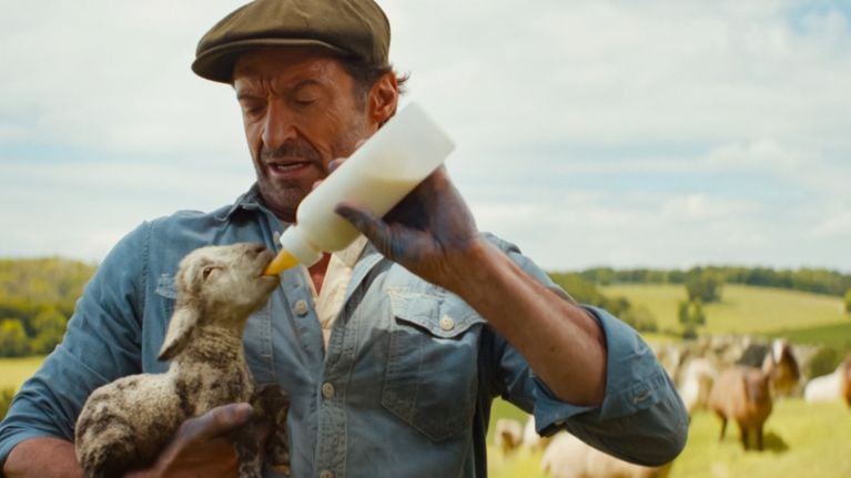 Hugh Jackman dressed as George Hardy in The Sheep Detectives, feeding a baby lamb from a bottle