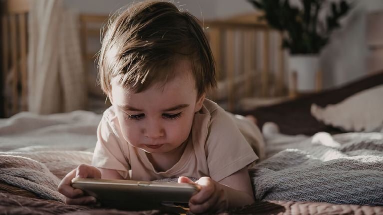 A focused toddler lying on a bed while holding and looking at a smartphone screen.