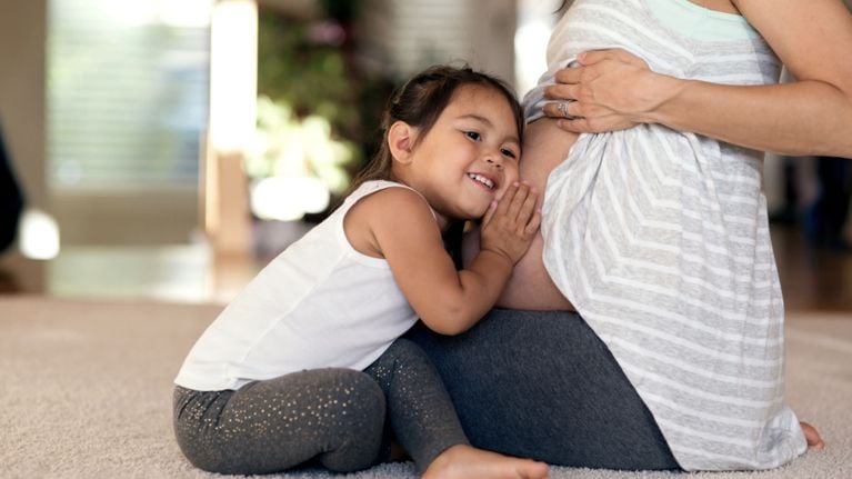 Young child leans their head against a pregnant parent’s belly while sitting on the floor, smiling up at the bump.
