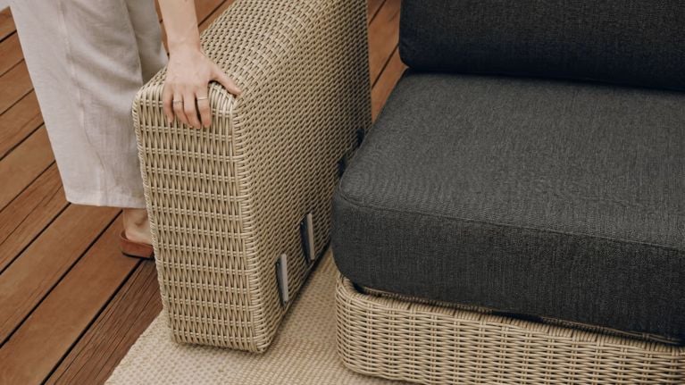 Close-up of a light wicker patio chair with a dark cushion as a person grips the armrest on a wood deck.