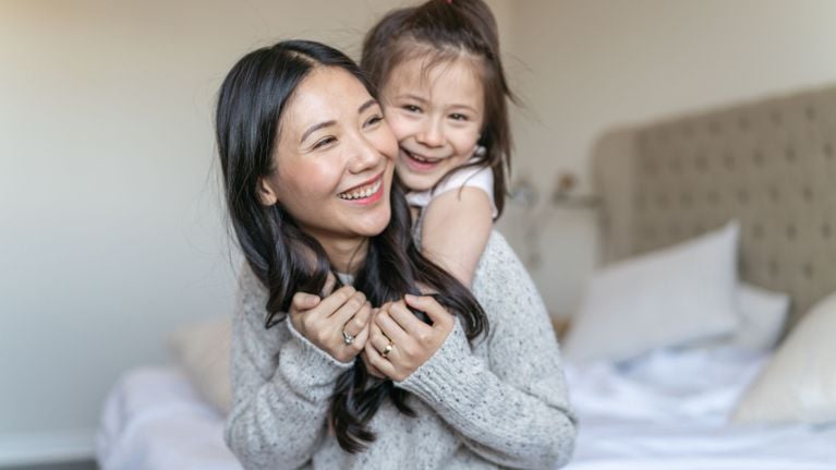 Pregnant parent smiles while giving a young child a piggyback ride in a softly lit bedroom.