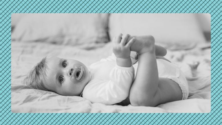 Black-and-white photo of an 8-month-old baby lying on a bed, holding one foot with both hands and looking toward the camera with a pacifier in their mouth, set against a blue patterned background.