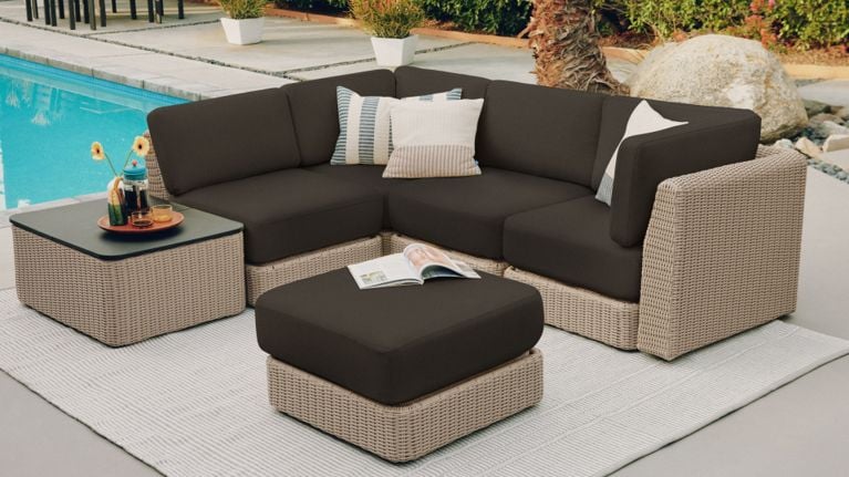 Light wicker outdoor sectional with dark cushions, a matching ottoman and side table, styled with pillows and drinks beside a pool.