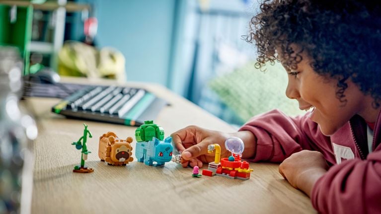 Child plays with small LEGO Pokémon figures on a table, including Bulbasaur and Bidoof, beside a tiny build with a clear dome.