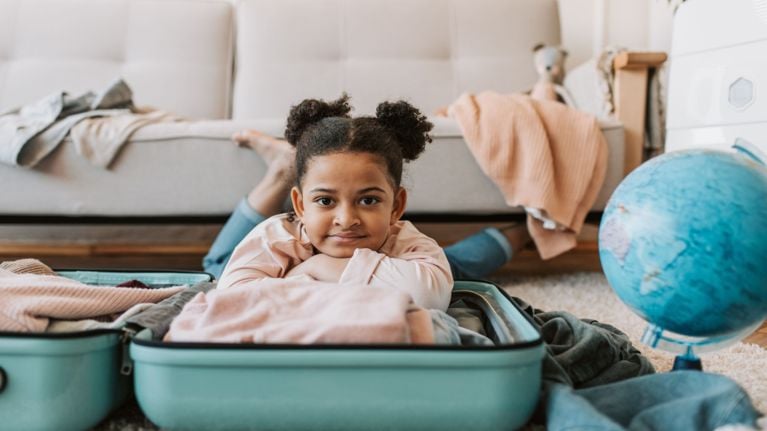 A child lies behind an open suitcase on the floor, looking at the camera, with clothes spread around and a globe nearby, suggesting family trip planning.