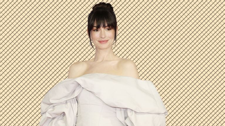 Anne Hathaway poses in an off-the-shoulder pale gown with dramatic puffed sleeves, her hair in a high bun, in front of a cream grid-pattern backdrop.