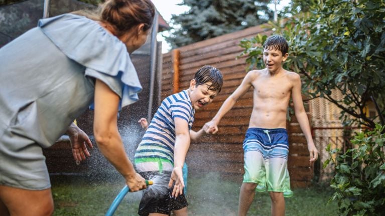 A woman sprays two boys in swim trunks with a hose in a backyard.