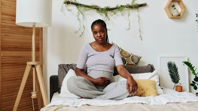 Pregnant Black woman sitting cross-legged on a bed in a bright bedroom, one hand resting on her belly, looking calmly at the camera.