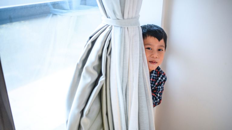 A young boy peering out from behind a bundle of curtains
