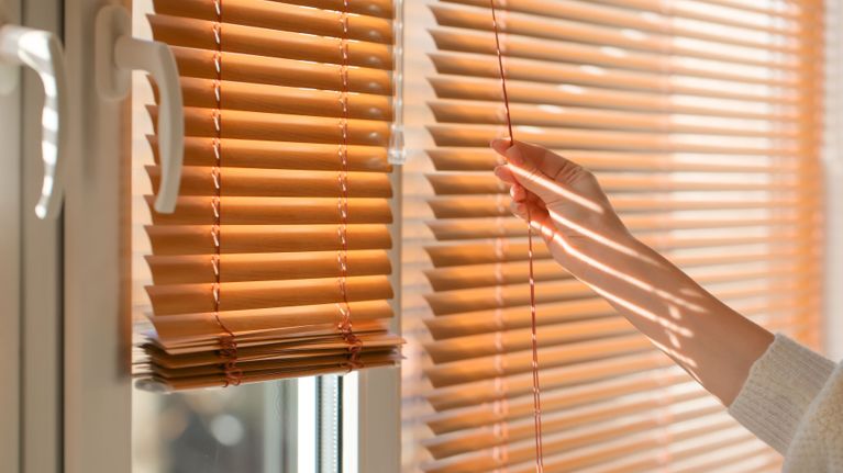 Hand pulling the cord on wooden window blinds beside a sunlit window.