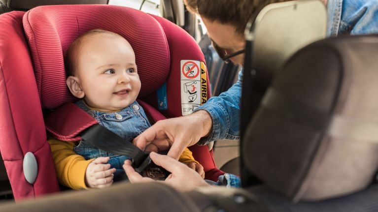 Parent fastening a smiling baby into a car seat in the back seat of a vehicle.
