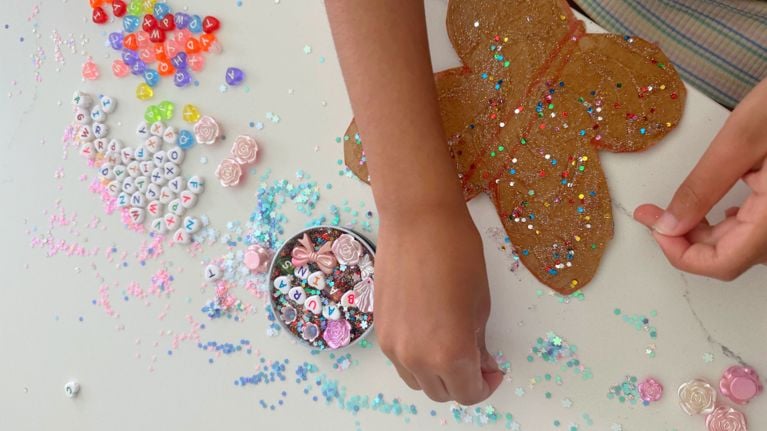 Overhead view of two children’s hands decorating glittery gingerbread-person shapes on a white table scattered with pastel letter beads, sequins and pink charms.