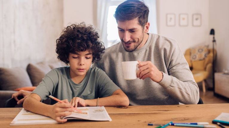 Parent sits beside a child at a table while the child works on a math worksheet, offering calm support at home.
