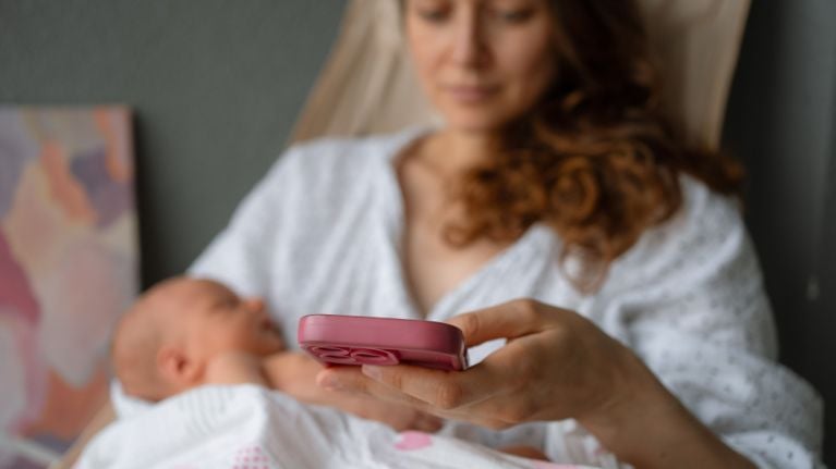 A mother holding a newborn in one arm while looking at a smartphone.