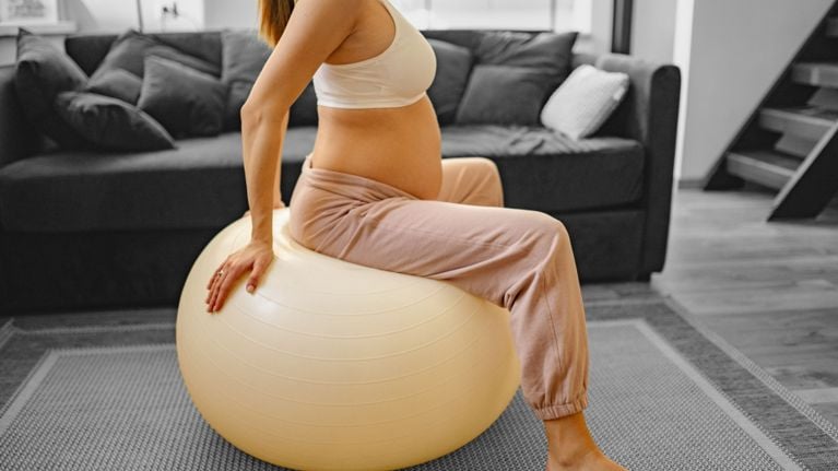 Pregnant person sitting sideways on a beige exercise ball in a living room, shown from the shoulders down, wearing a white tank top and pink joggers.