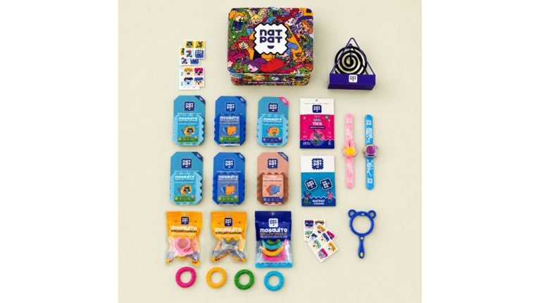 Flat lay of NatPat bug-repellent products and accessories for kids, including sticker packs, wristbands, charms and tins, arranged on a pale background.