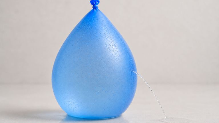 Blue water balloon leaking a thin stream of water from the side against a plain light background, illustrating urinary leakage during pregnancy.