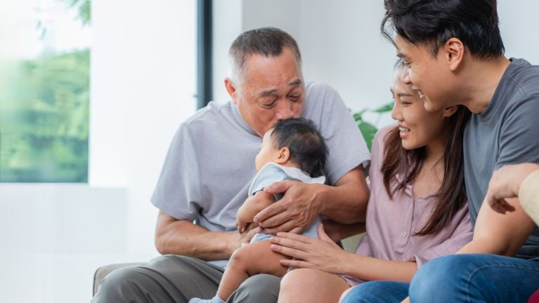 An elderly man kisses the top of a baby's head while a younger couple smiles next to them on a sofa
