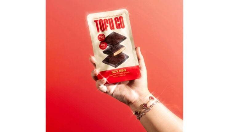 Hand holding a package of Tofu Go soy BBQ savoury protein snack against a bright red background.