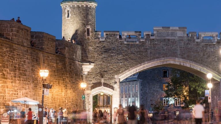 Historic stone gate in Old Quebec lit at dusk, with pedestrians and streetlights glowing below the arch.