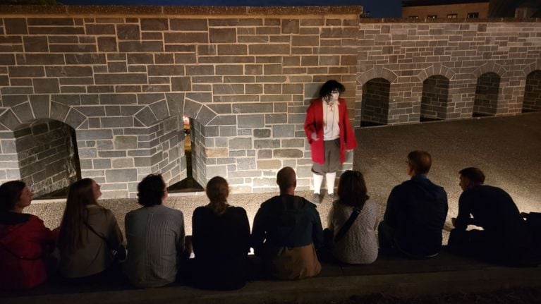 Costumed performer speaking to a seated audience at night beside a stone fortification wall.
