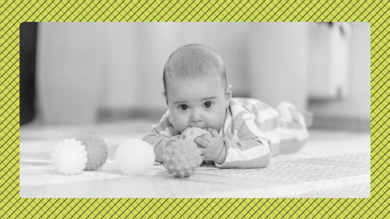 Black-and-white photo of a baby lying on their tummy on the floor, chewing a toy and looking at the camera, with three textured balls in the foreground and a yellow patterned border around the image.