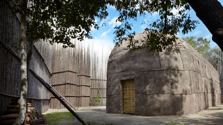 Traditional Indigenous-style dwelling and tall wooden palisade at an outdoor historic site, framed by trees.