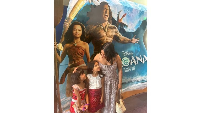A woman and two children stand in front of a large Moana live-action movie display featuring Moana and Maui. The poster includes the text “Disney Moana” and “In Theatres July 10.”