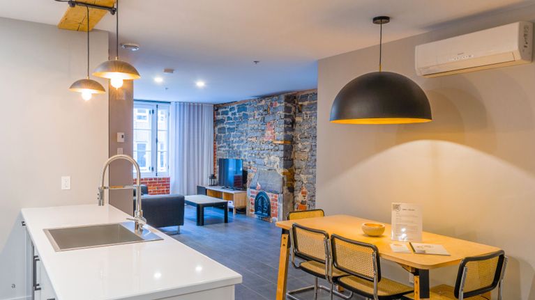 Modern loft-style apartment with a white kitchen island, dining table, exposed stone wall and small living area near a window.